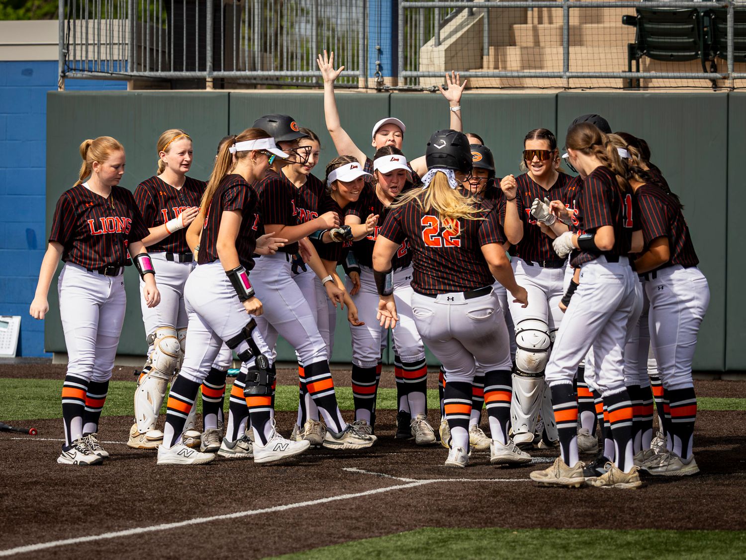 Softball players in maroon and white uniforms celebrate and gather around home plate to greet a teammate after a home run.