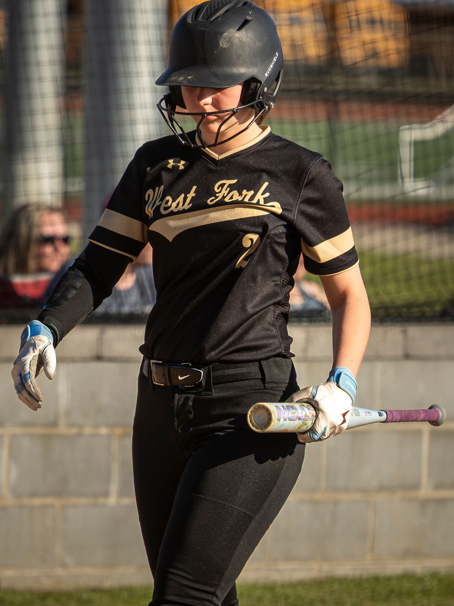 Softball player in a black West Fork jersey and batting helmet walks with a bat in hand along a fence near the dugout.