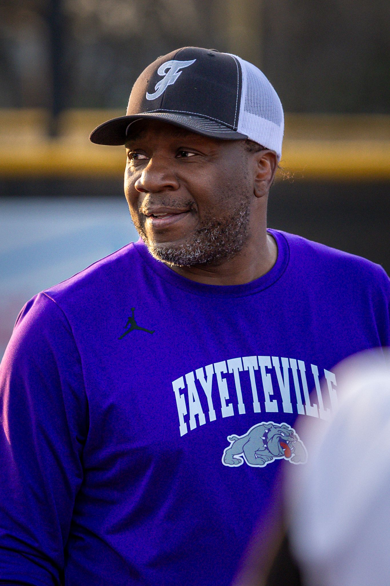 Smiling coach in a black Fayetteville cap and purple Bulldogs shirt looks off to the side at a softball field.