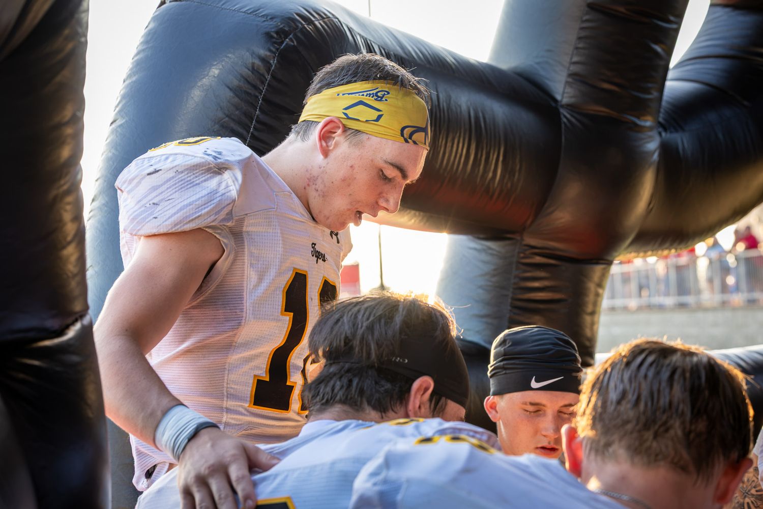 Football player in a white jersey and yellow headband huddles with teammates near an inflatable tunnel before a game.