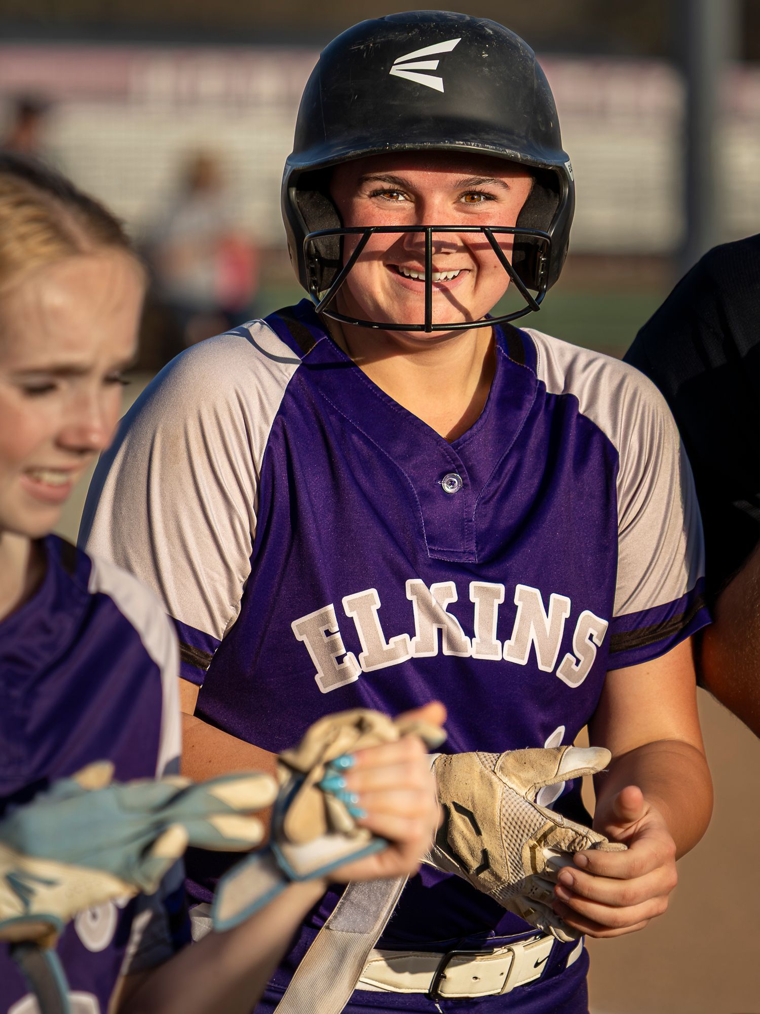 A smiling softball player in a purple Elkins jersey and black batting helmet with a face guard stands holding her gloves during a game.