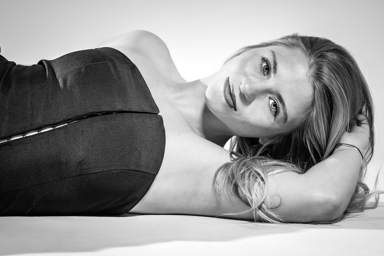 Black and white photo of a blonde woman lying down, wearing a black top and jeans, looking at the camera.