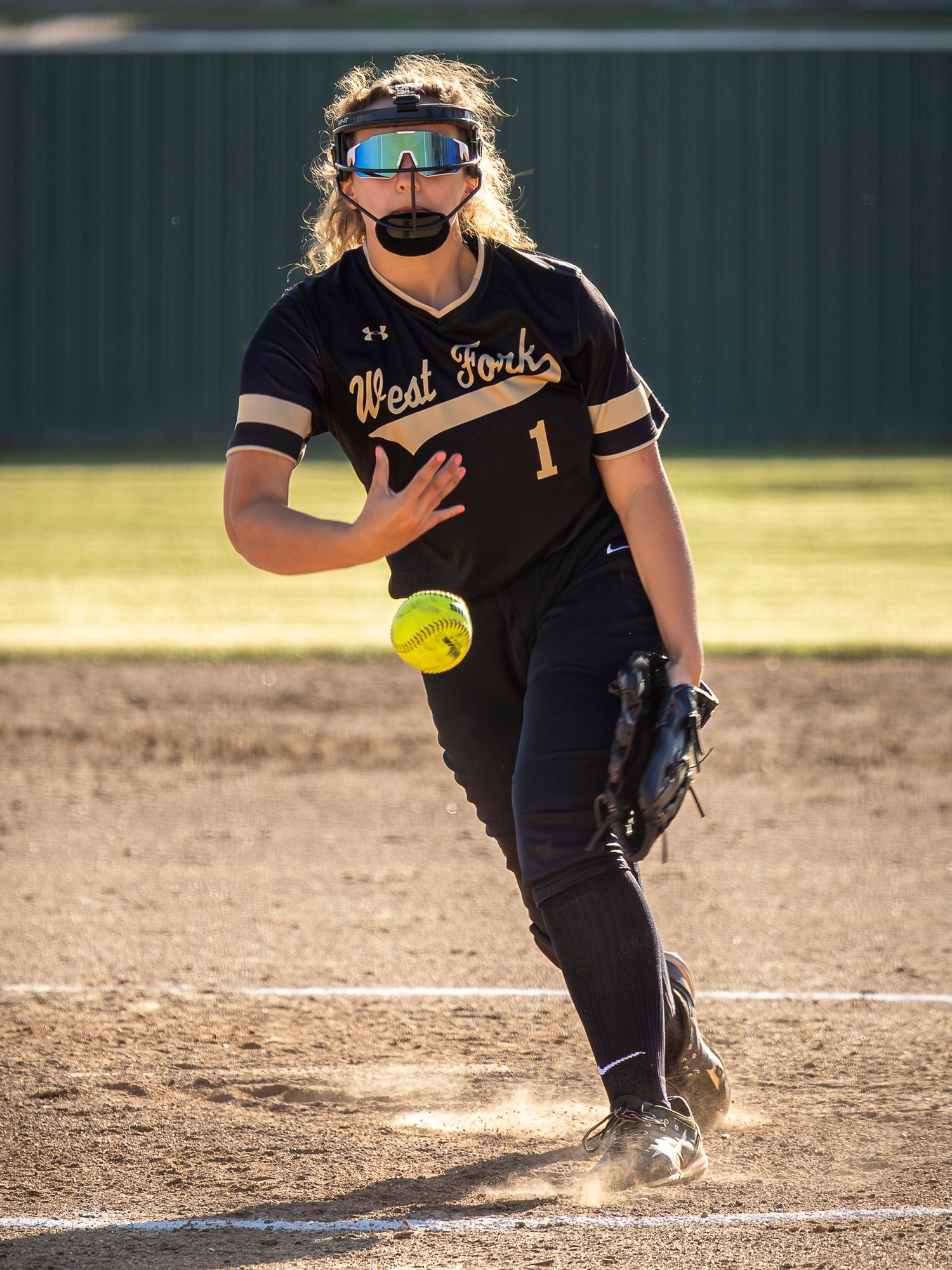 Softball pitcher in black uniform releasing a yellow ball during a game on a dirt field in bright sunlight.