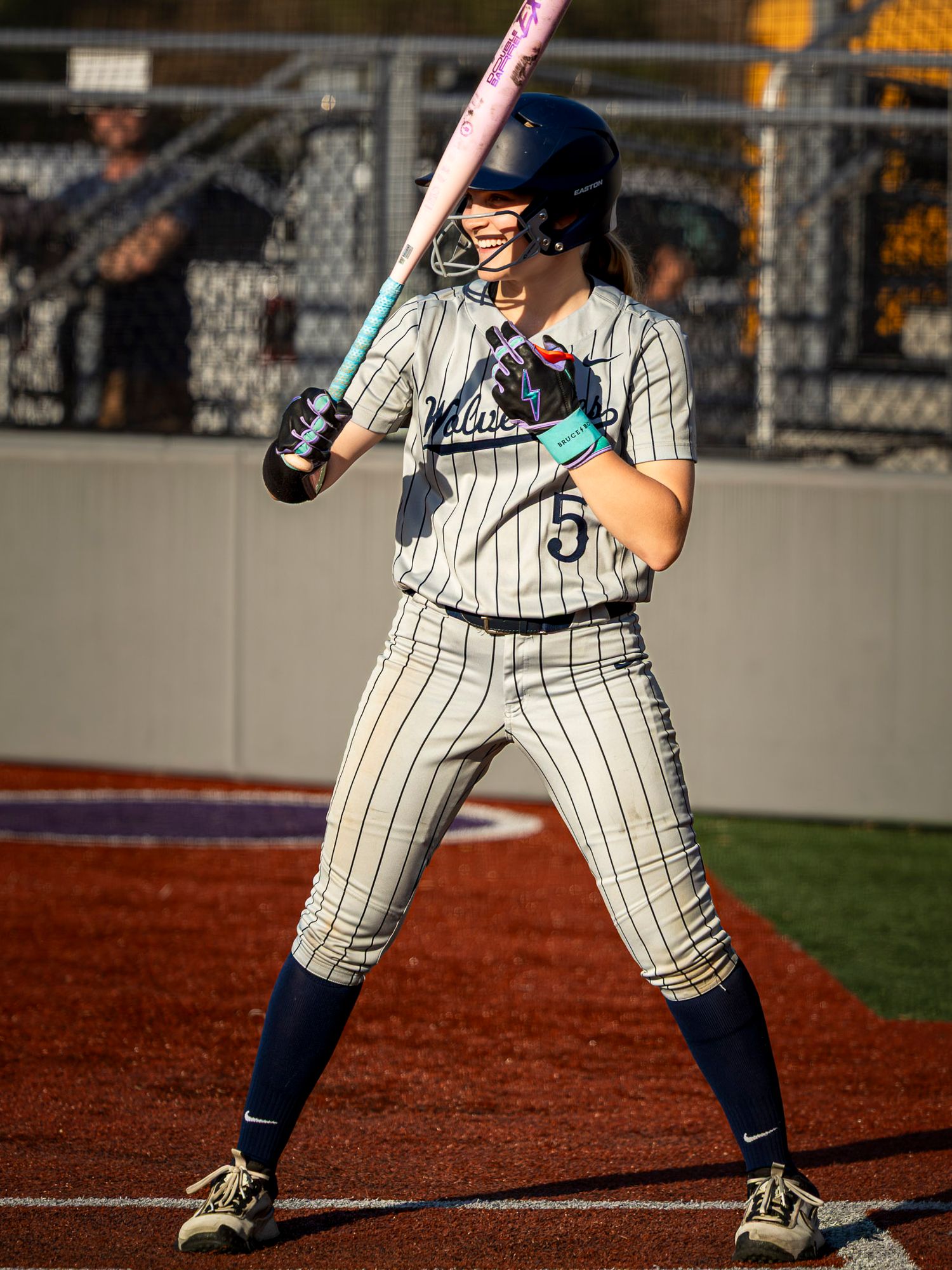 Softball player wearing number 5 stands at bat in pinstripe uniform, holding pink bat in ready stance on field.