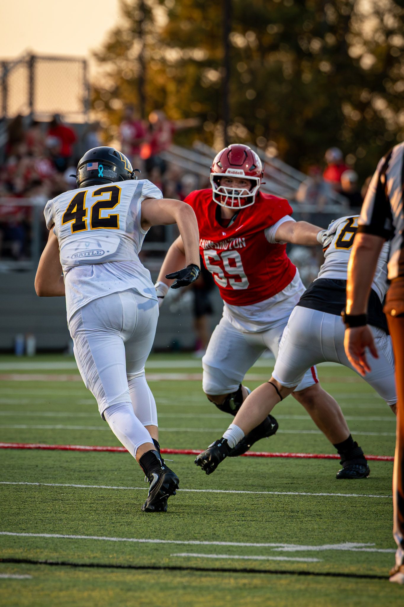 Football player in white jersey number 42 rushes past a red-jerseyed opponent number 59 during a high school game at sunset.