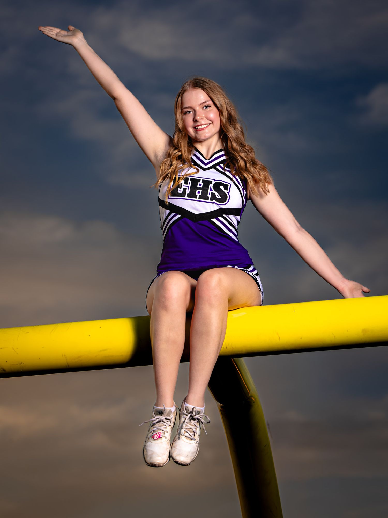 Smiling cheerleader in a purple EHS uniform sits on a yellow football goalpost with one arm raised against a dramatic evening sky.