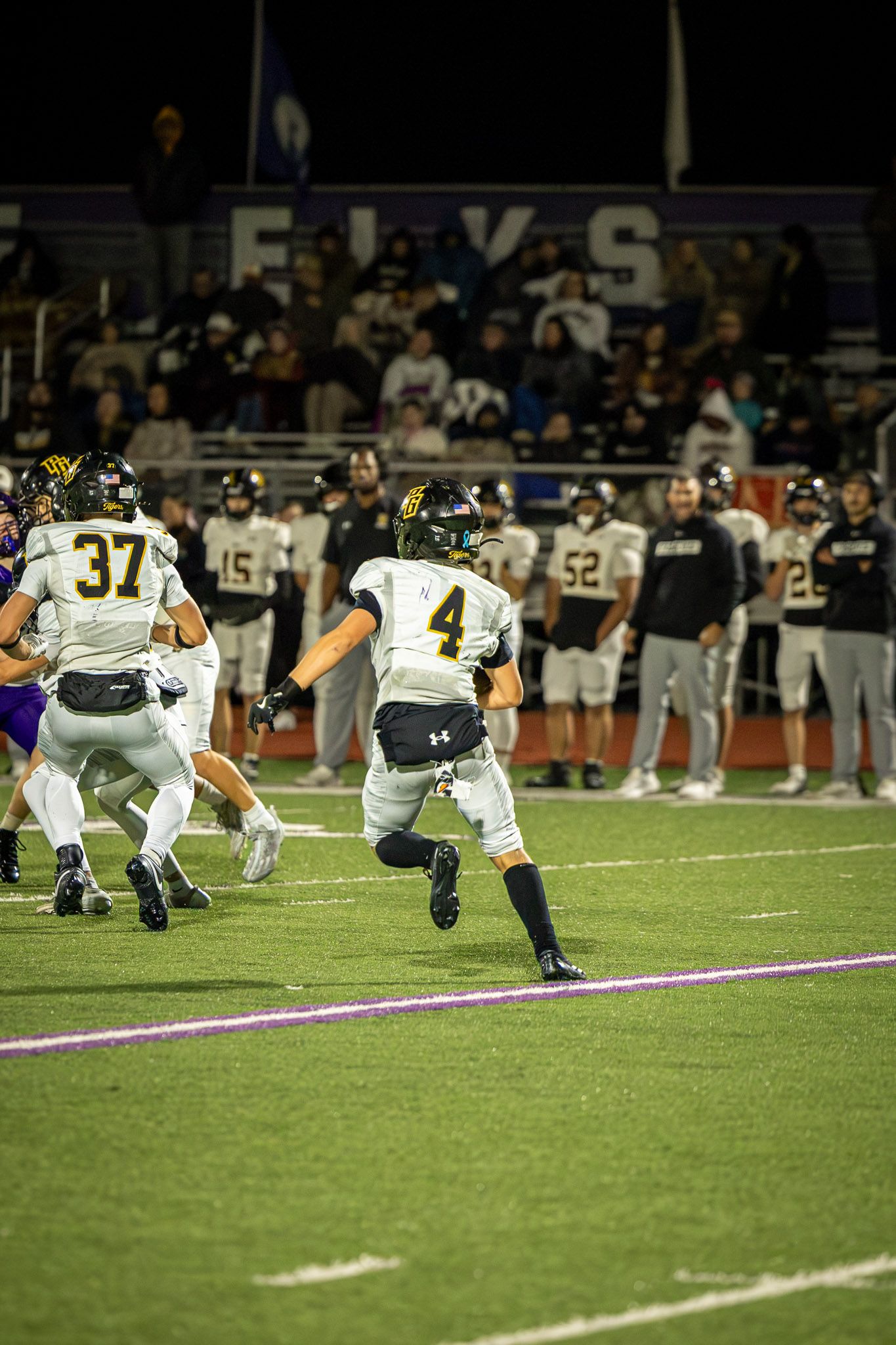 High school football player wearing number 4 runs downfield during a night game while teammates and sideline coaches watch from the field.