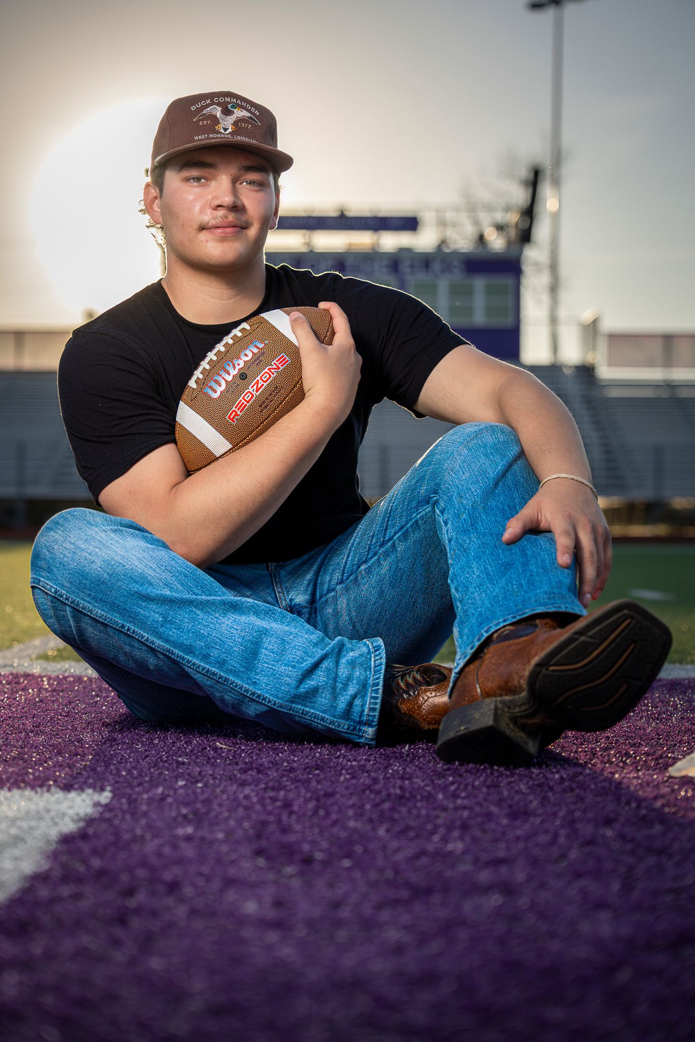 A young man in a baseball cap sits on a purple field, holding a football in his hands.