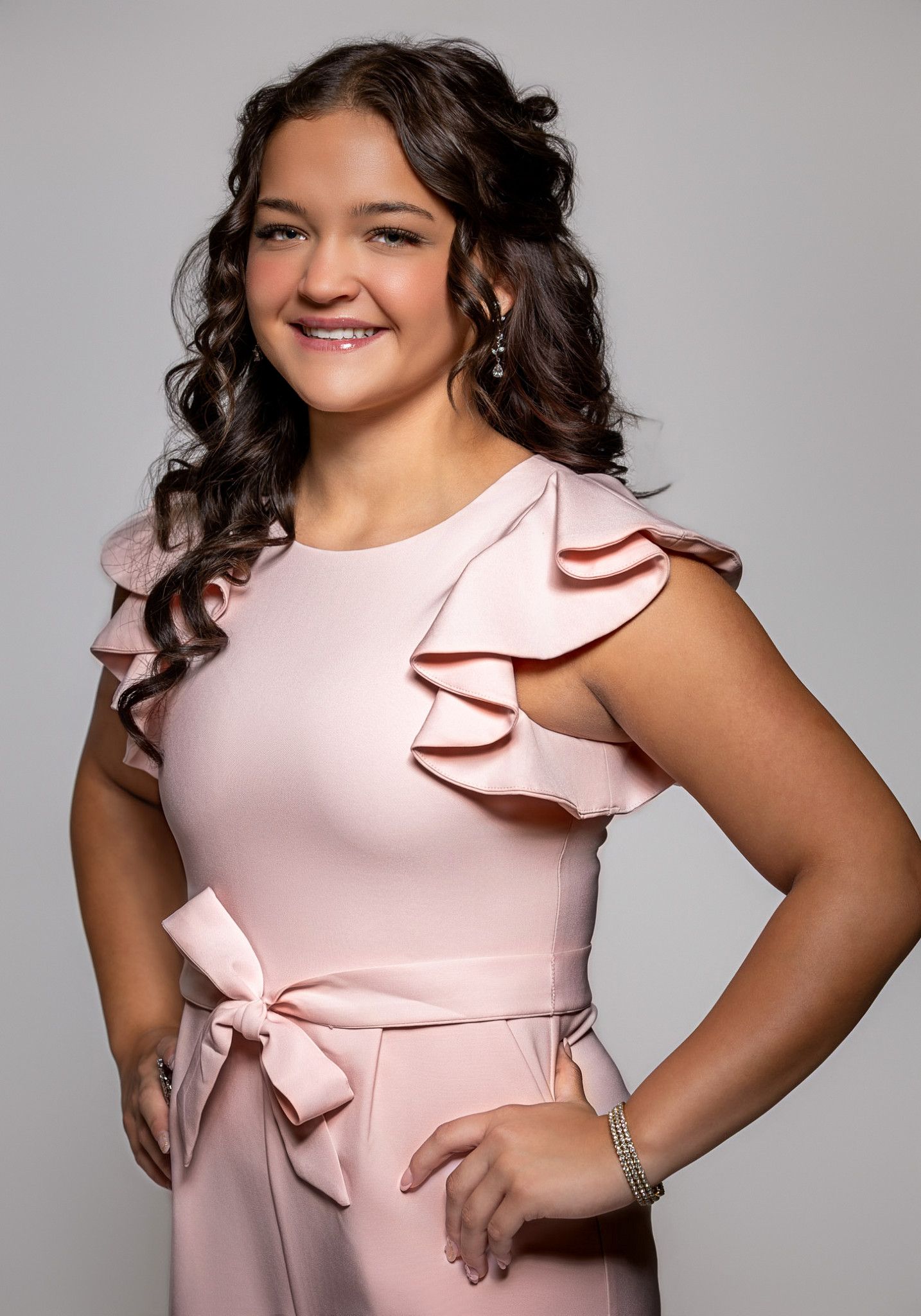 Young woman smiling in a pink ruffled dress with bow detail, posing confidently against a gray background.