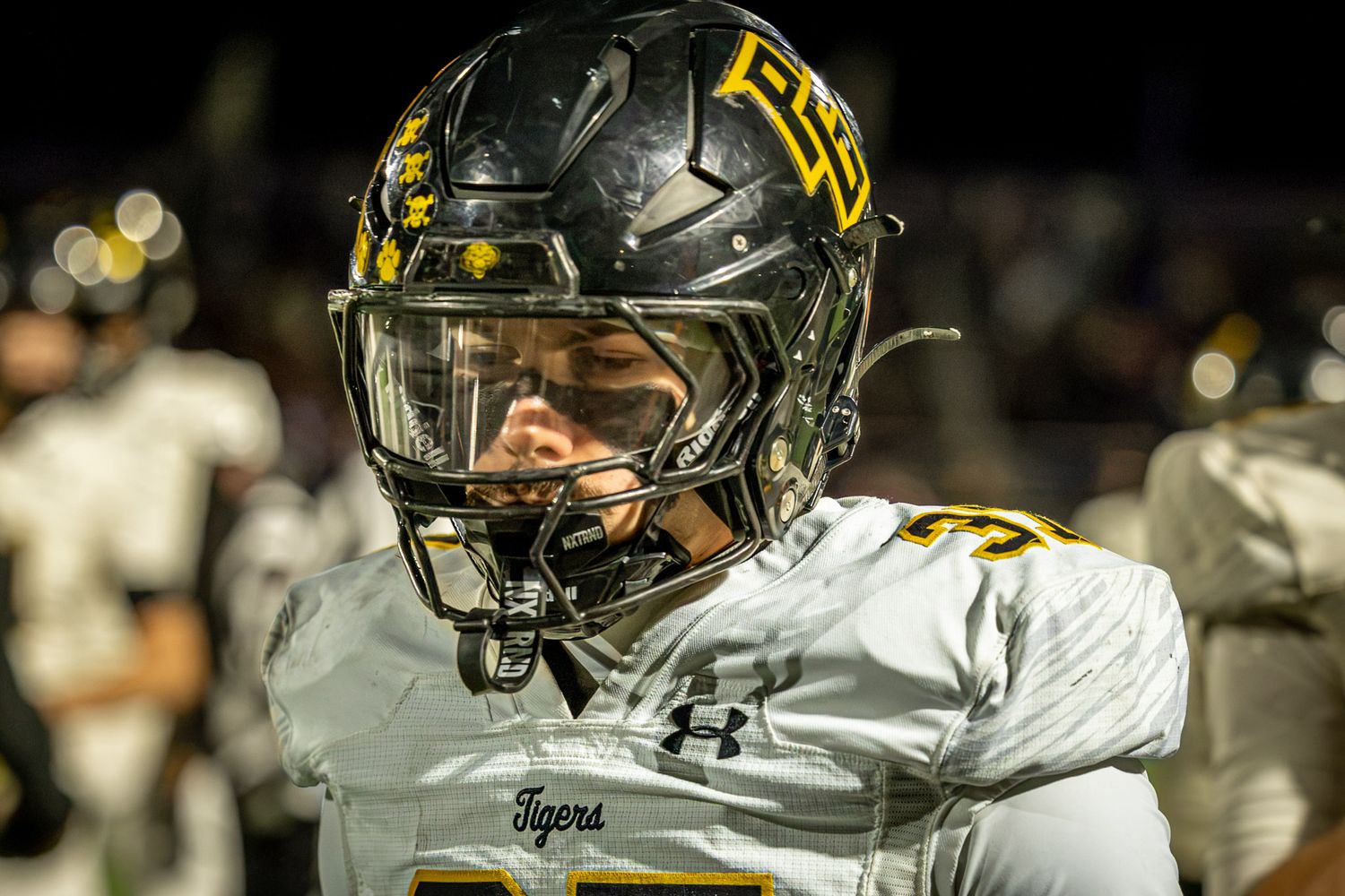 Football player in a black helmet with gold lettering and a white Tigers jersey stands on the field during a night game.