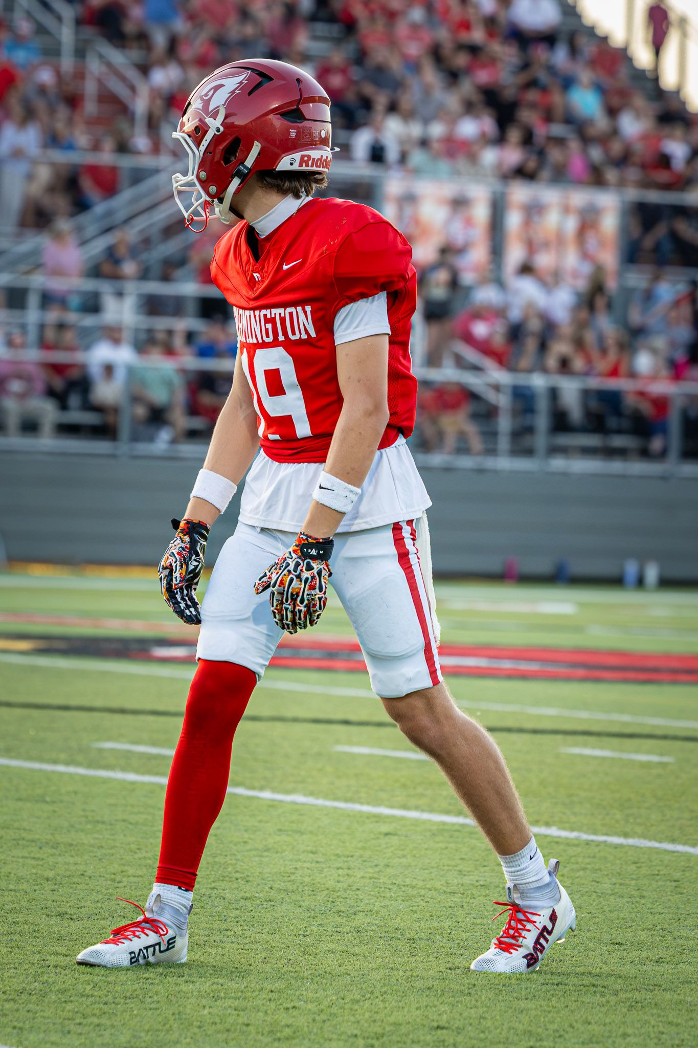 High school football player in a red number 19 jersey, helmet, and gloves stands ready on the field during a game with fans in the stands.