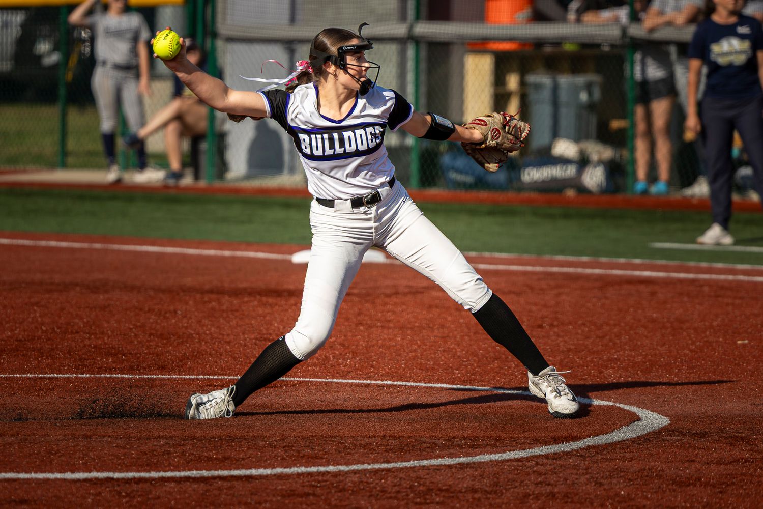 Bulldogs softball pitcher in a white uniform winds up to throw a pitch from the circle on a red clay field.