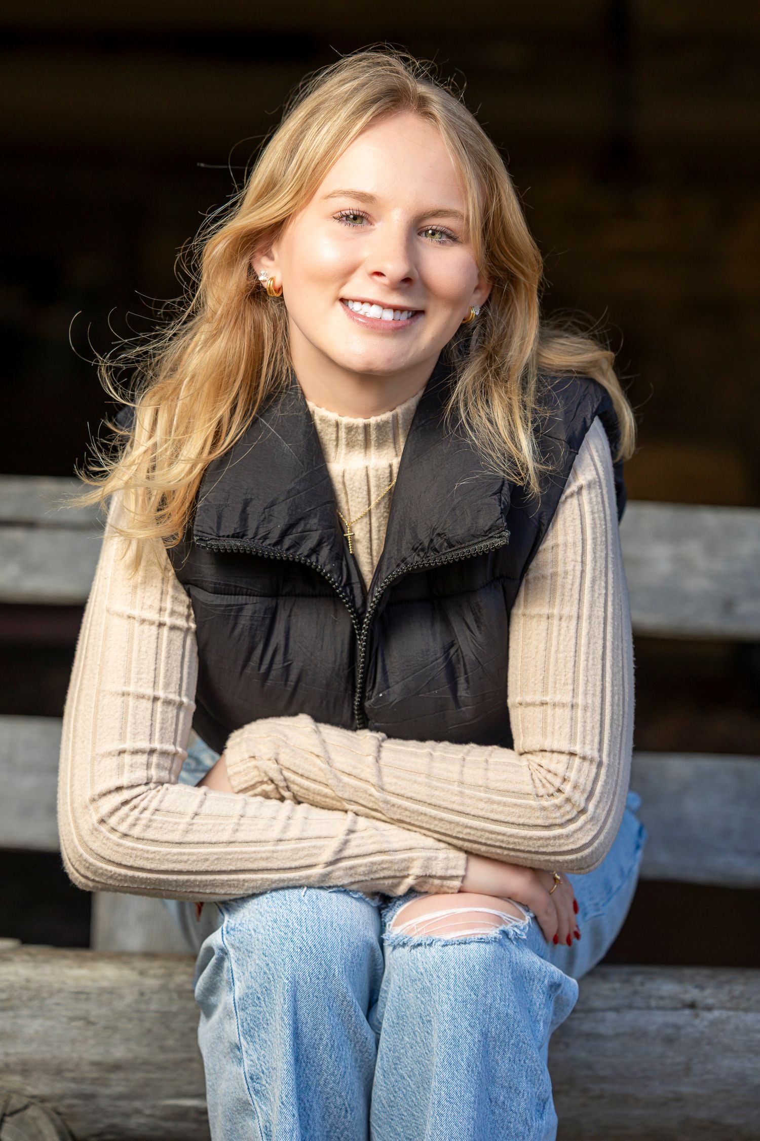 Smiling young blonde woman in black vest and jeans sitting on rustic wooden fence in warm sunlight.