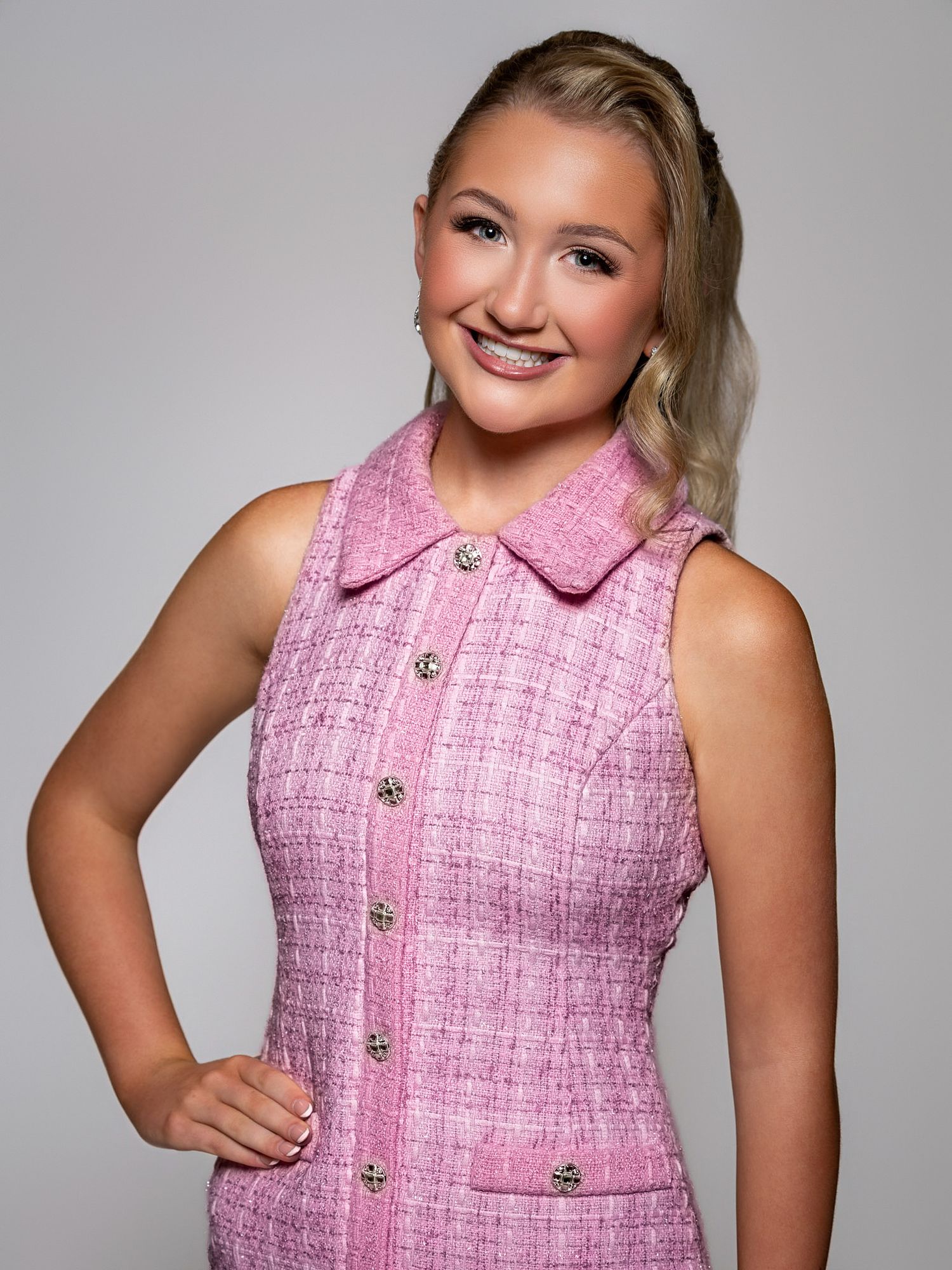Young woman smiling in a pink tweed sleeveless dress with silver buttons, posing confidently against a gray background.