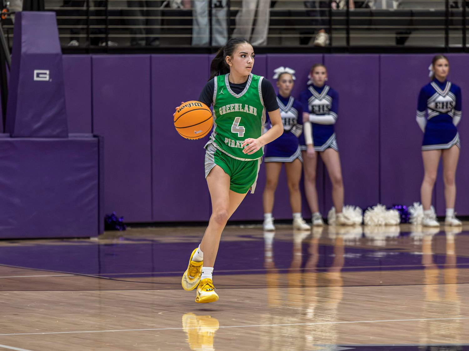 Basketball player in green jersey dribbles down court during game while cheerleaders watch in background.