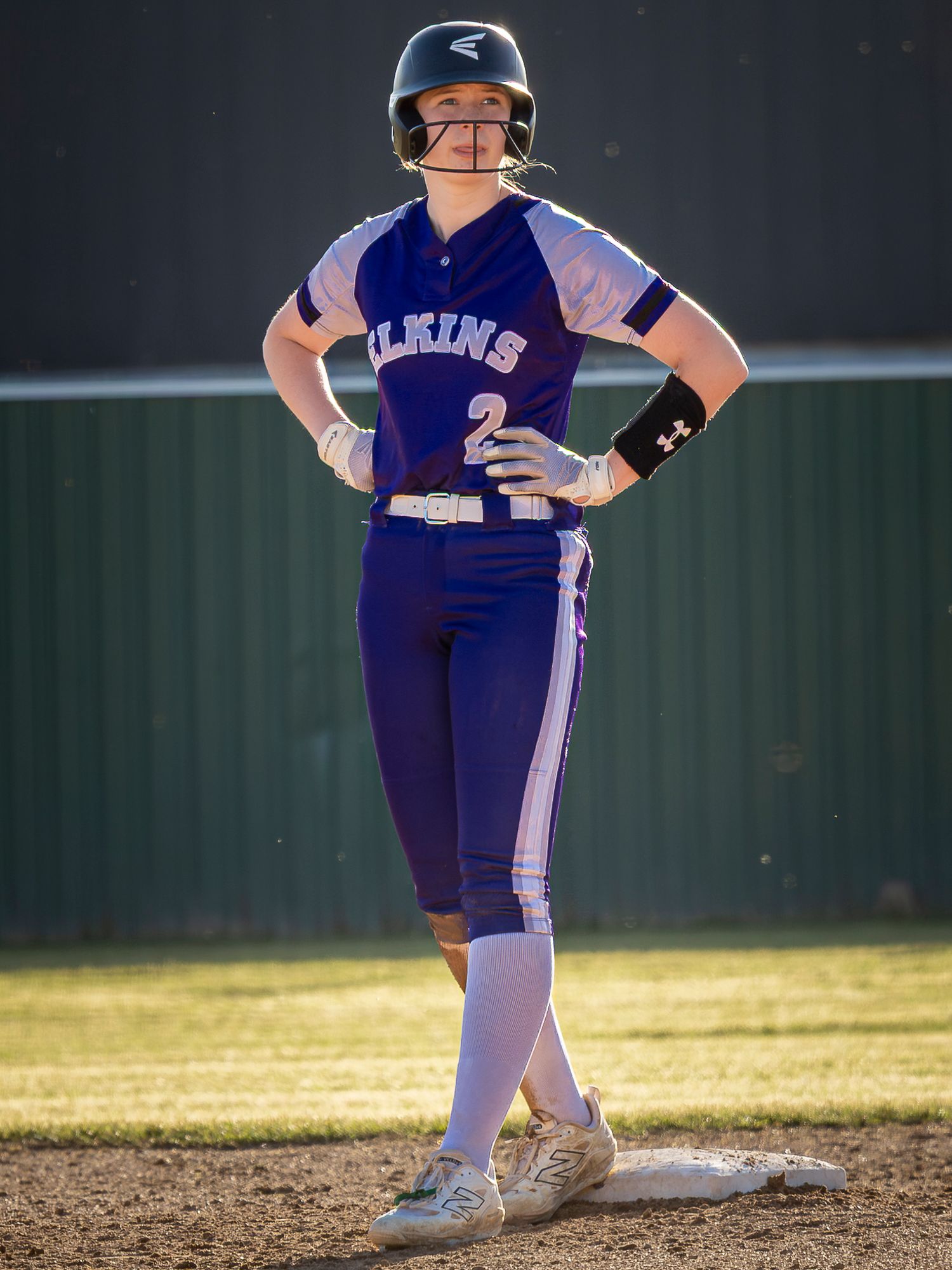 Elkins softball player in a purple number 2 uniform and batting helmet stands on a base with one foot on the bag during a game.