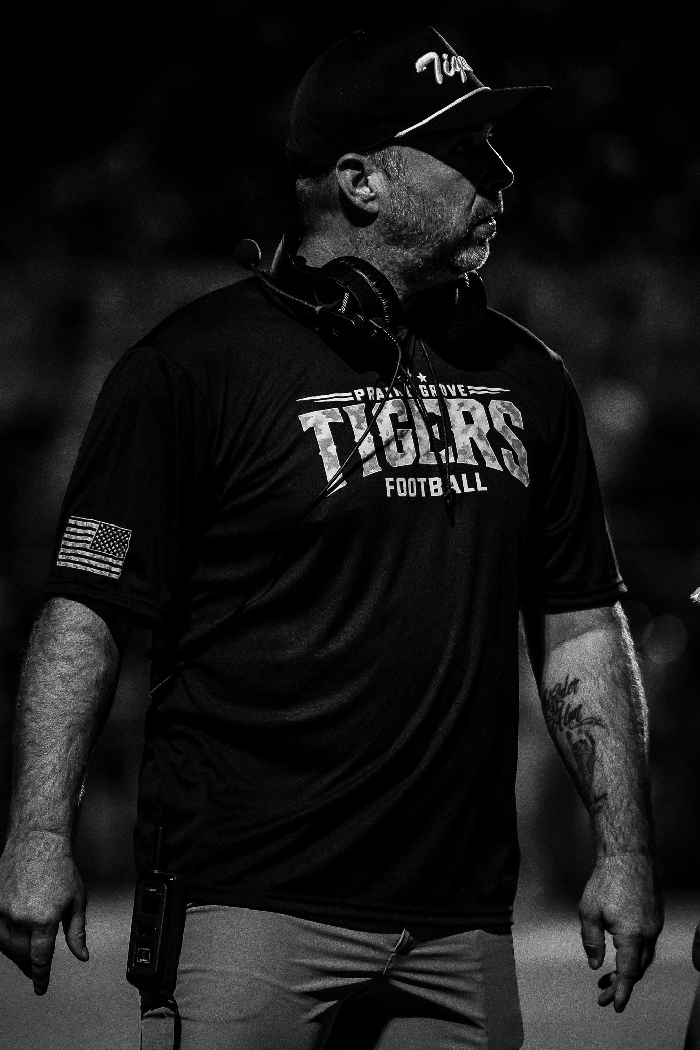 Football coach in a black Tigers Football shirt and headset looks to the side during a night game in black and white.