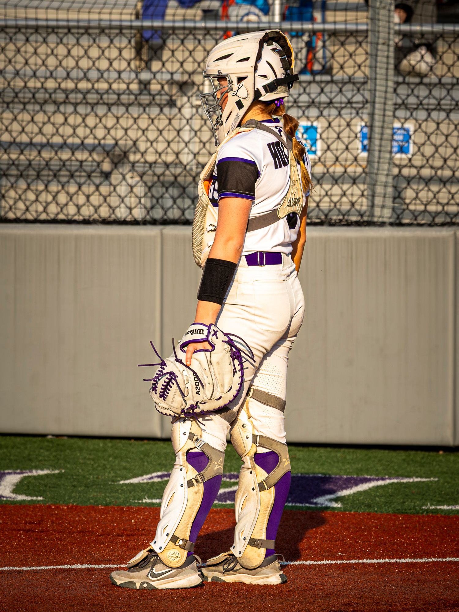 Softball catcher in a white helmet, leg guards and purple-trimmed uniform holds her mitt and waits behind home plate during a game.