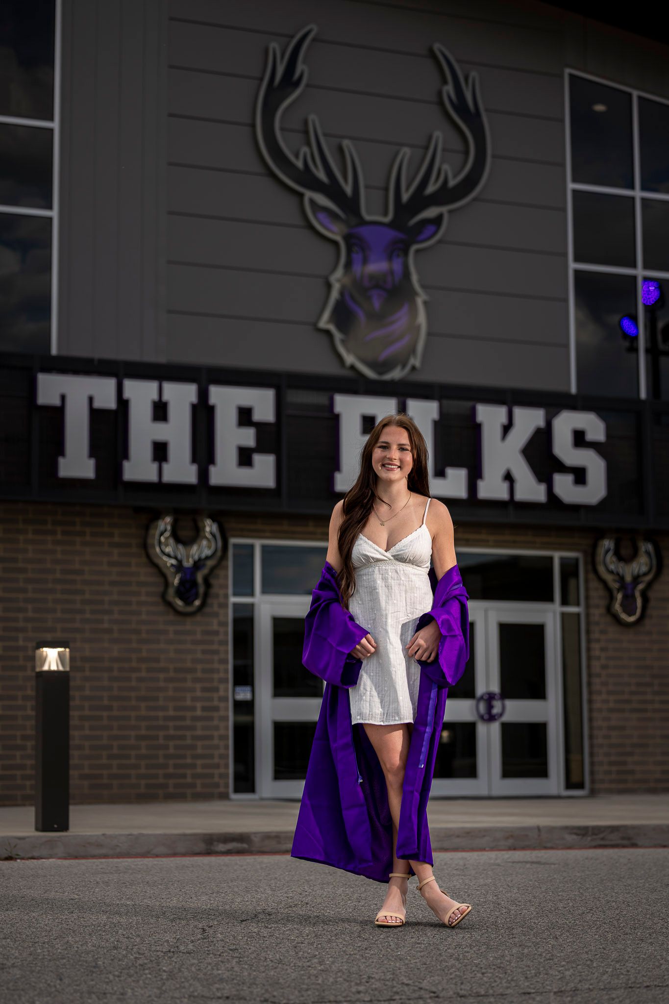 Elkins senior graduate in white dress and purple robe poses smiling in front of The Elks building entrance at night.