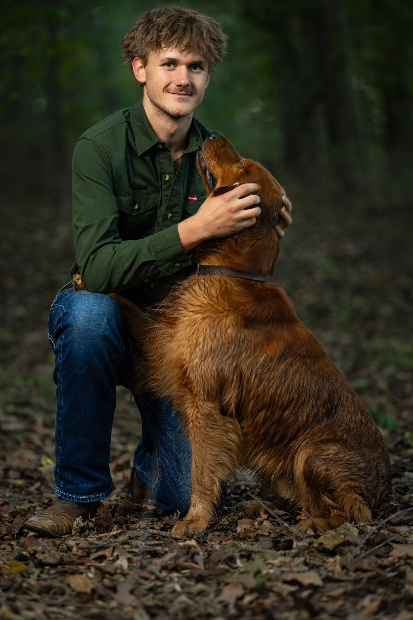 Young man in a green button-up shirt and jeans kneels in a leafy forest, gently petting a golden retriever gazing up at him.