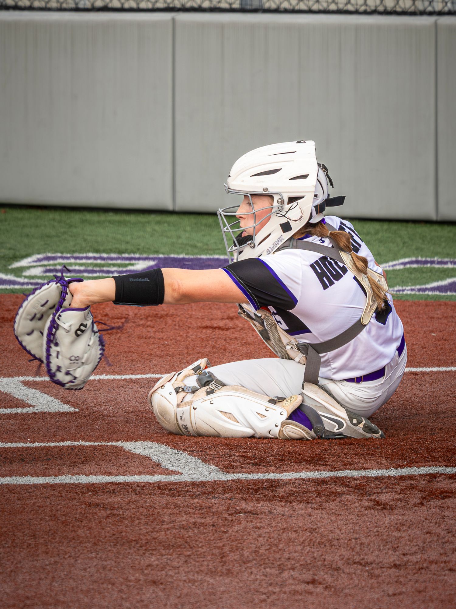 Softball catcher in a white helmet and purple jersey crouches behind home plate with her mitt extended to receive a pitch.