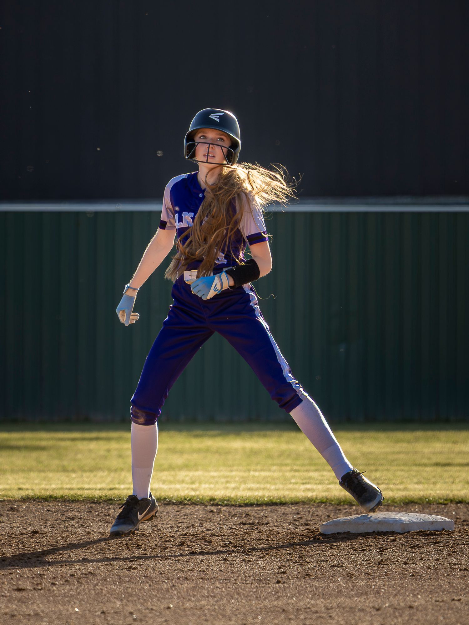 A softball baserunner in a purple uniform and batting helmet stands on second base with long hair flying during a game.