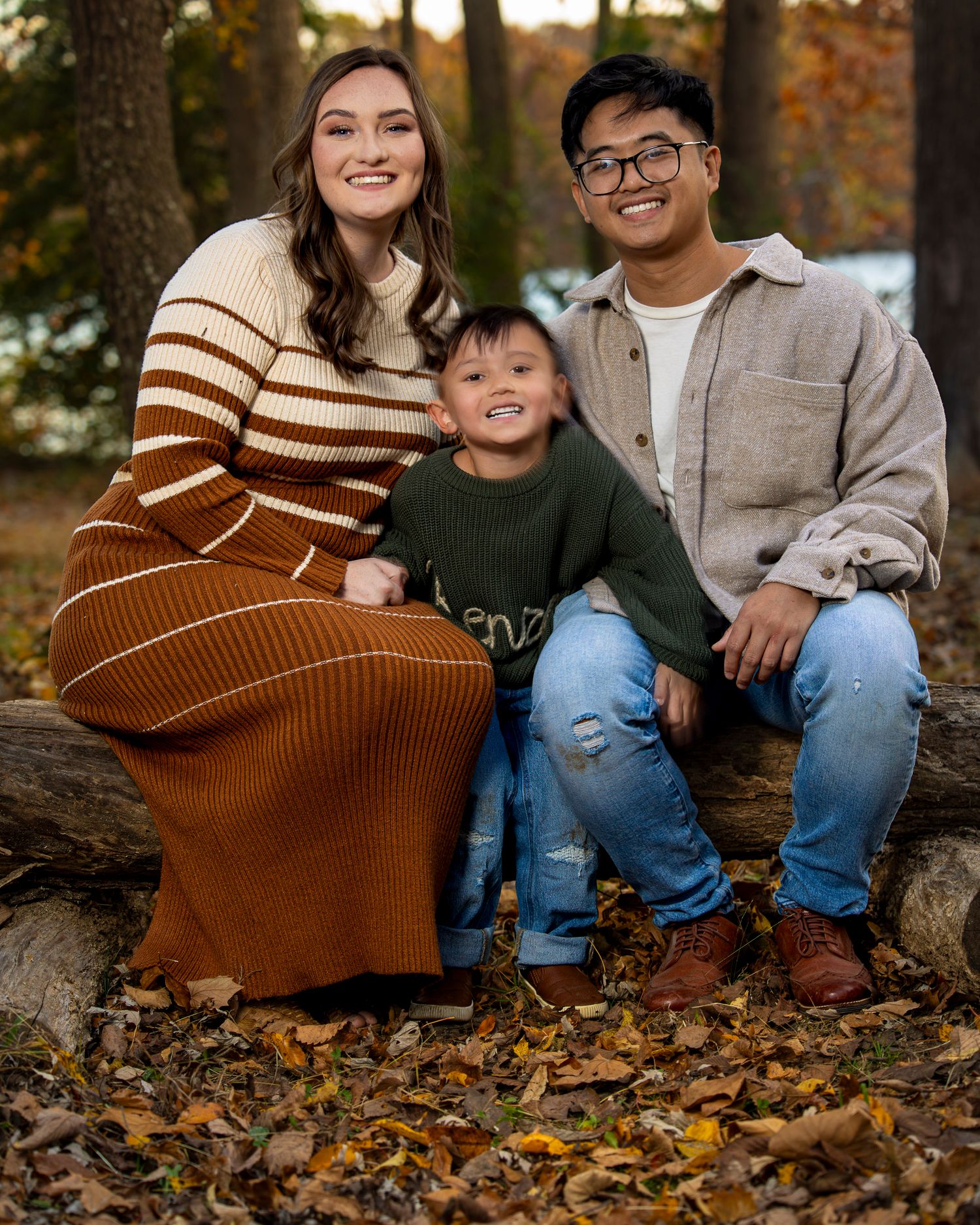 A family sits together on fallen logs surrounded by autumn leaves in a forest setting during a fall photo session.