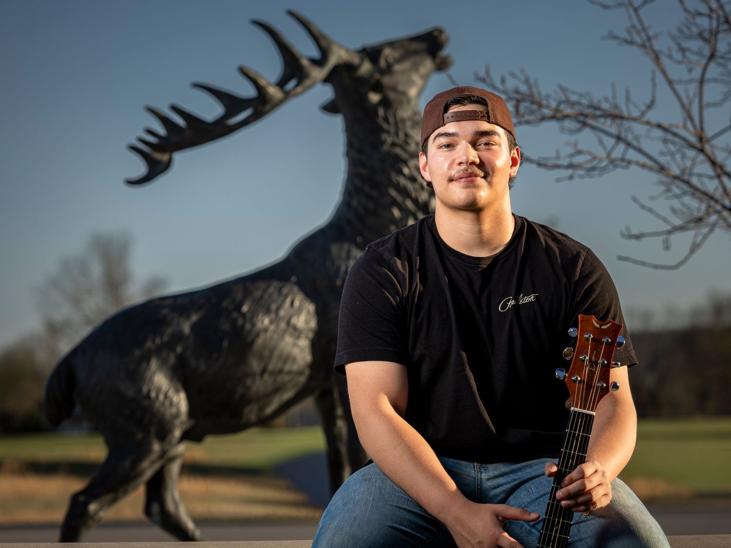 Young man holding a guitar poses in front of a large black elk statue outdoors at golden hour.