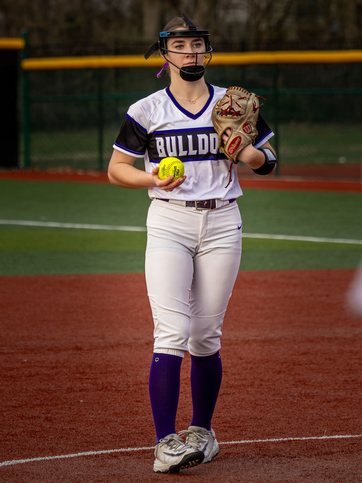 Bulldogs softball pitcher in a white and purple uniform and face guard holds a yellow softball and glove while standing on the infield dirt.
