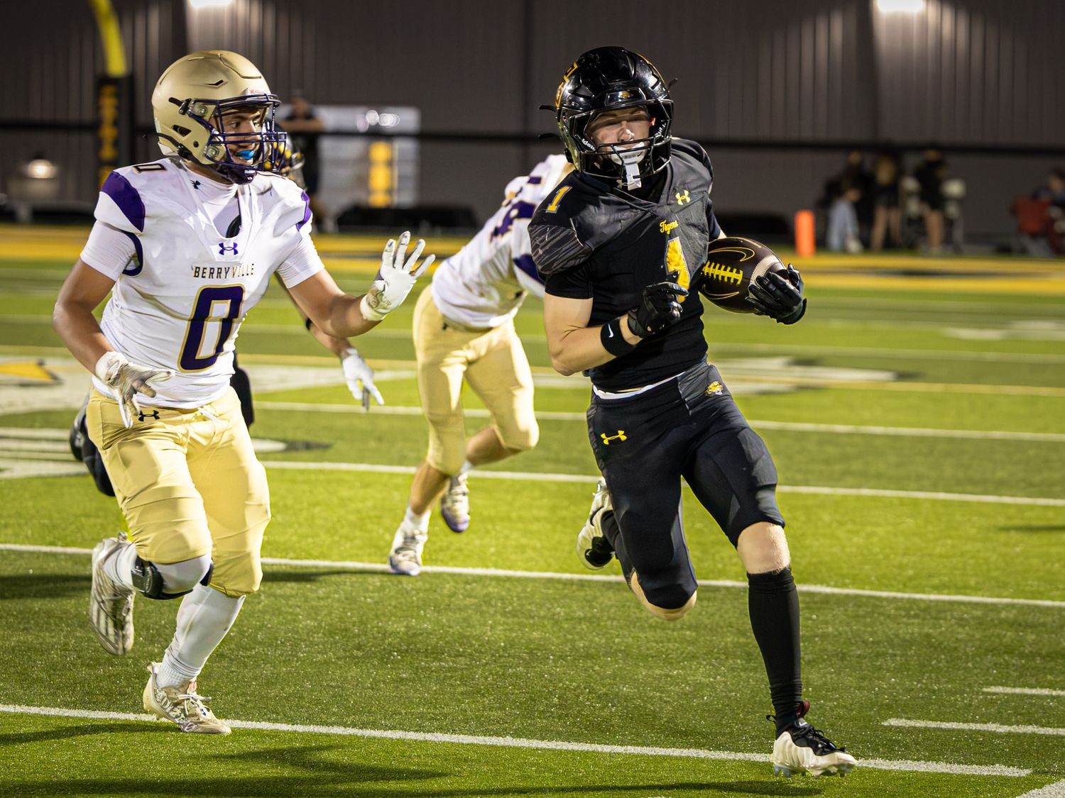A high school football player in a black uniform carries the ball downfield while a defender in a white and purple jersey chases him.