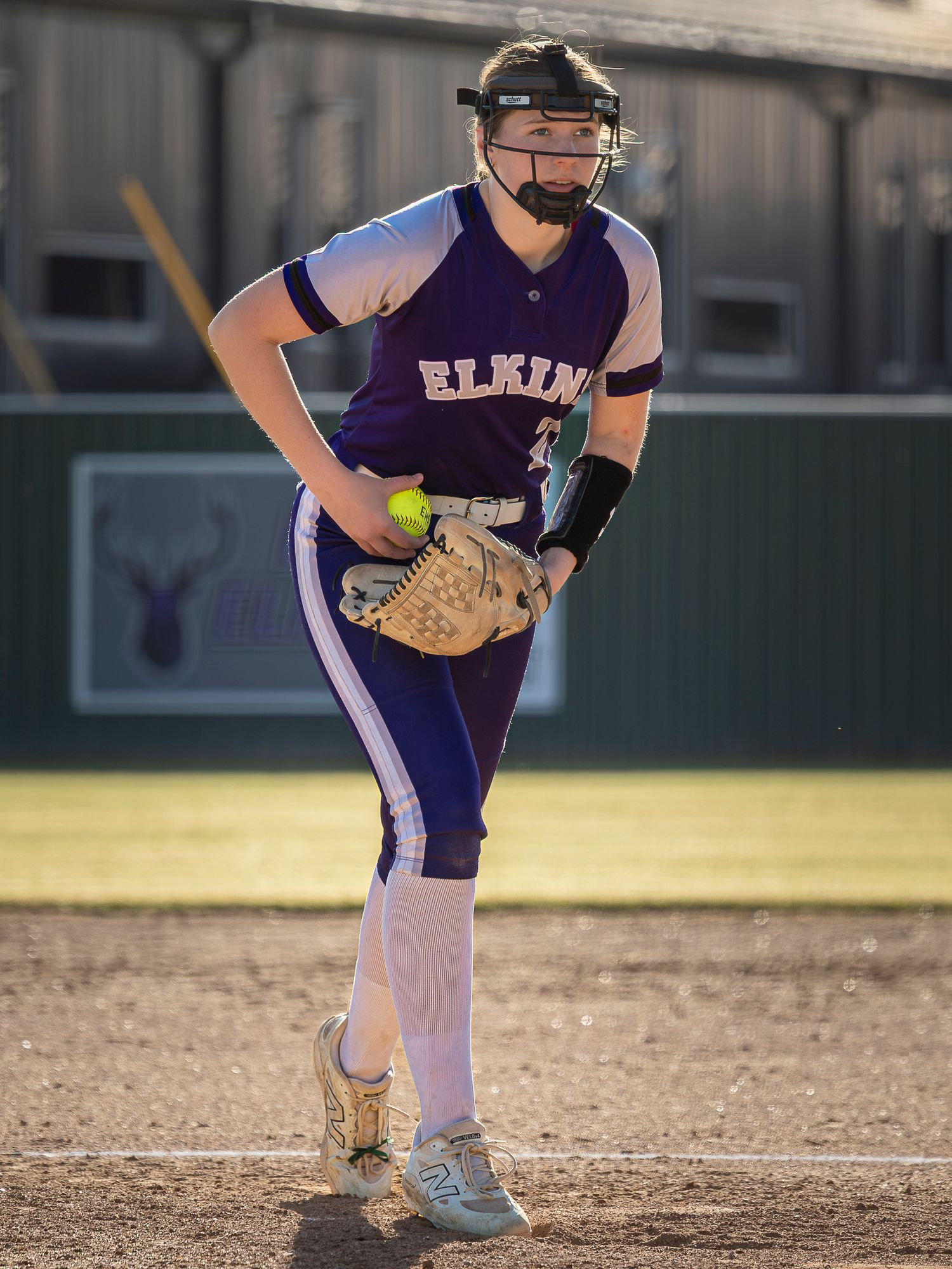Elkins softball pitcher in a purple jersey and face guard winds up with a yellow softball in her glove on a dirt field.