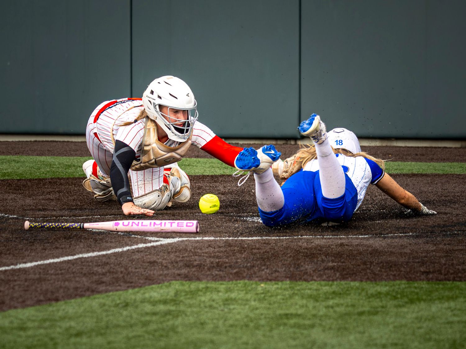 Softball catcher in white gear reaches to tag a sliding runner in blue at home plate as a yellow softball lies in the dirt nearby.