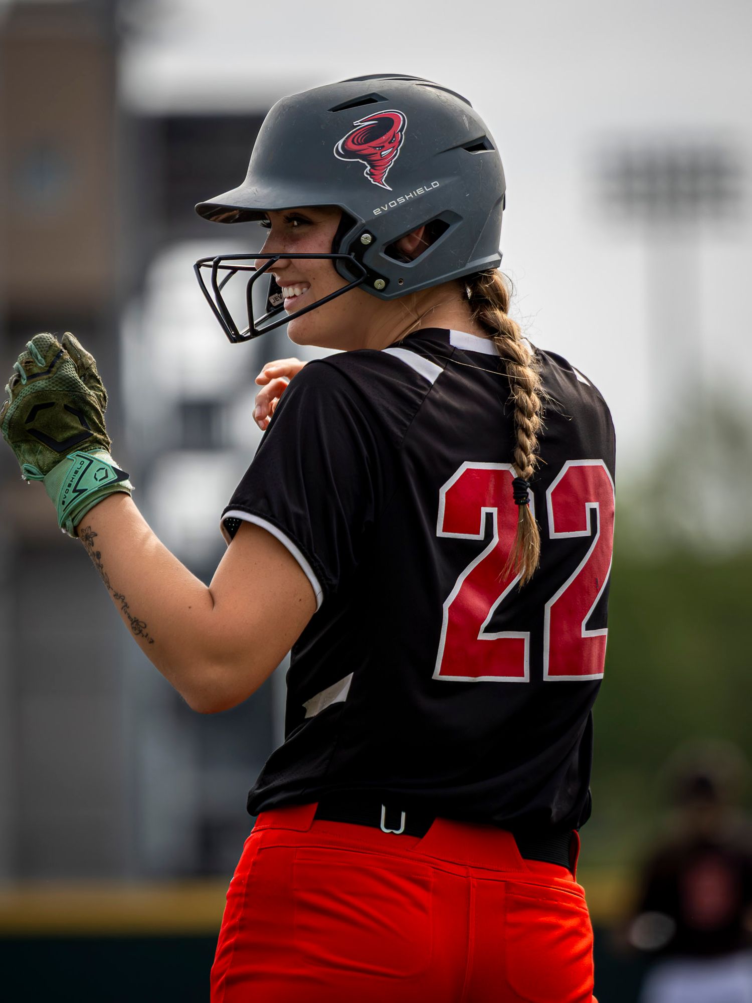 Smiling softball player in a black number 22 jersey, red pants and gray batting helmet with a face guard, wearing green batting gloves.
