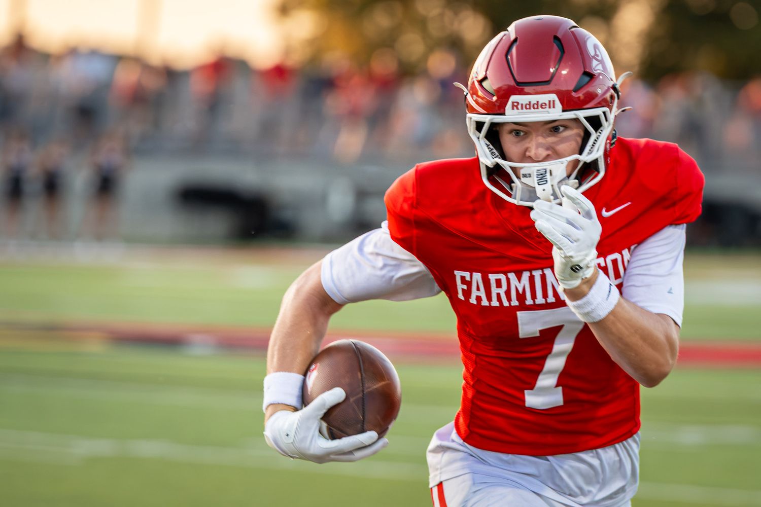 Football player in a red Farmington jersey with number 7 and helmet runs along the field while carrying the ball during a game.