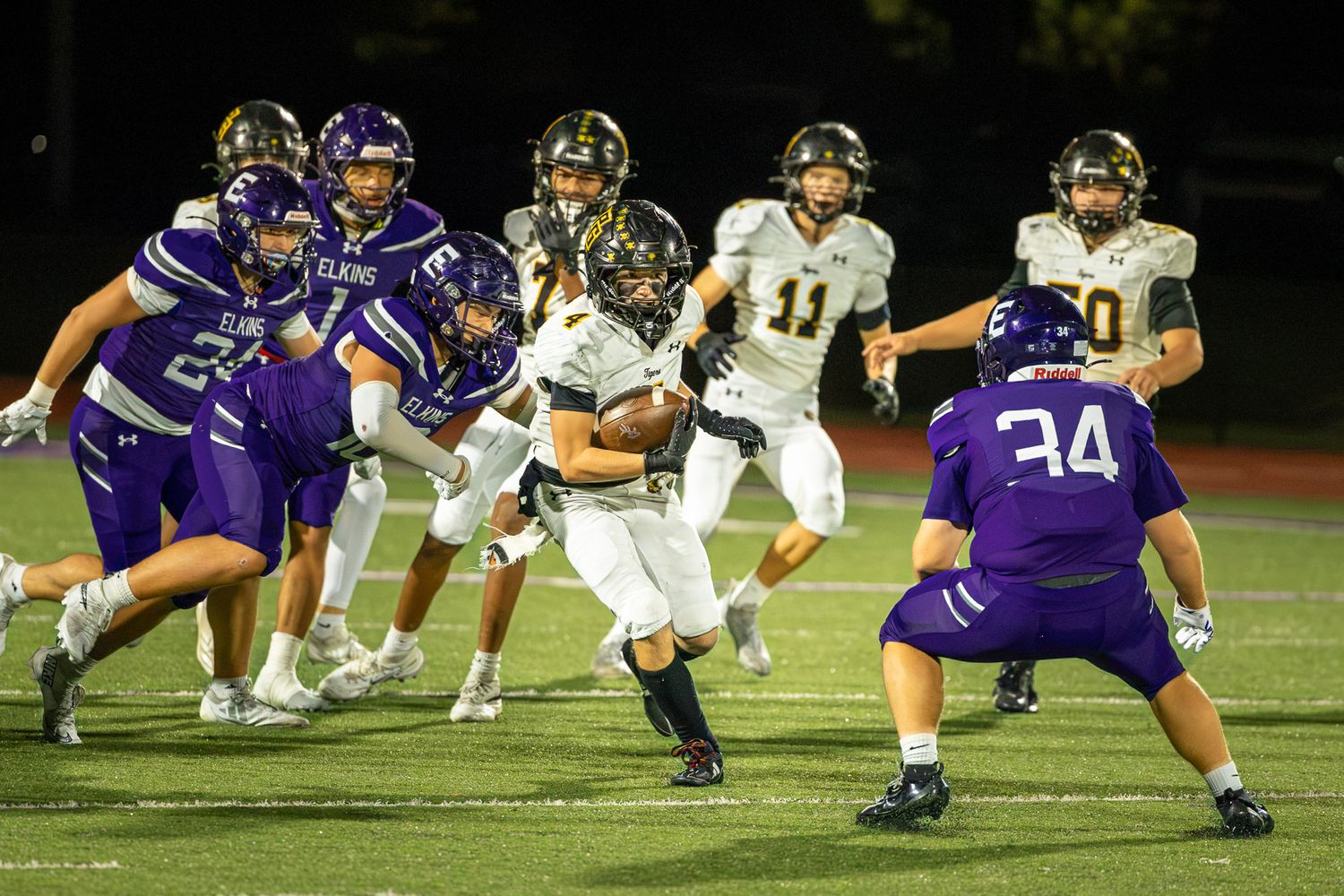 High school football player in a white jersey carries the ball past defenders in purple uniforms during a Friday night game.