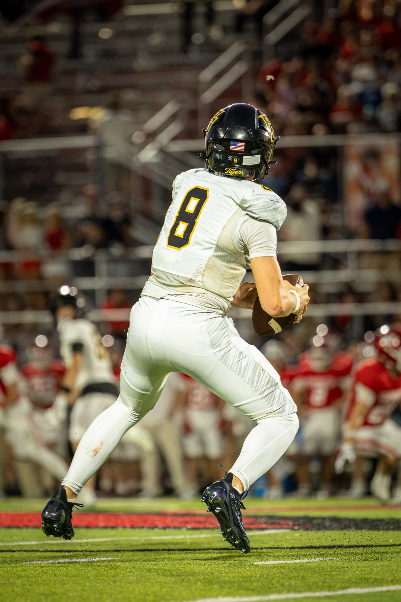 Football quarterback in white jersey number 8 and black helmet drops back to pass during a night game with fans in the stands behind him.