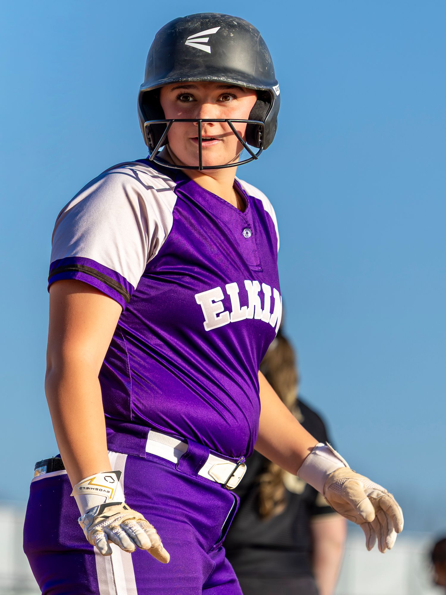 Smiling Elkins softball batter in a purple jersey, black helmet with face guard, and batting gloves looks over her shoulder.