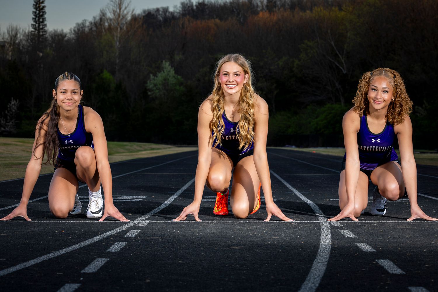 Three female track athletes in purple uniforms pose in starting position on an outdoor running track.