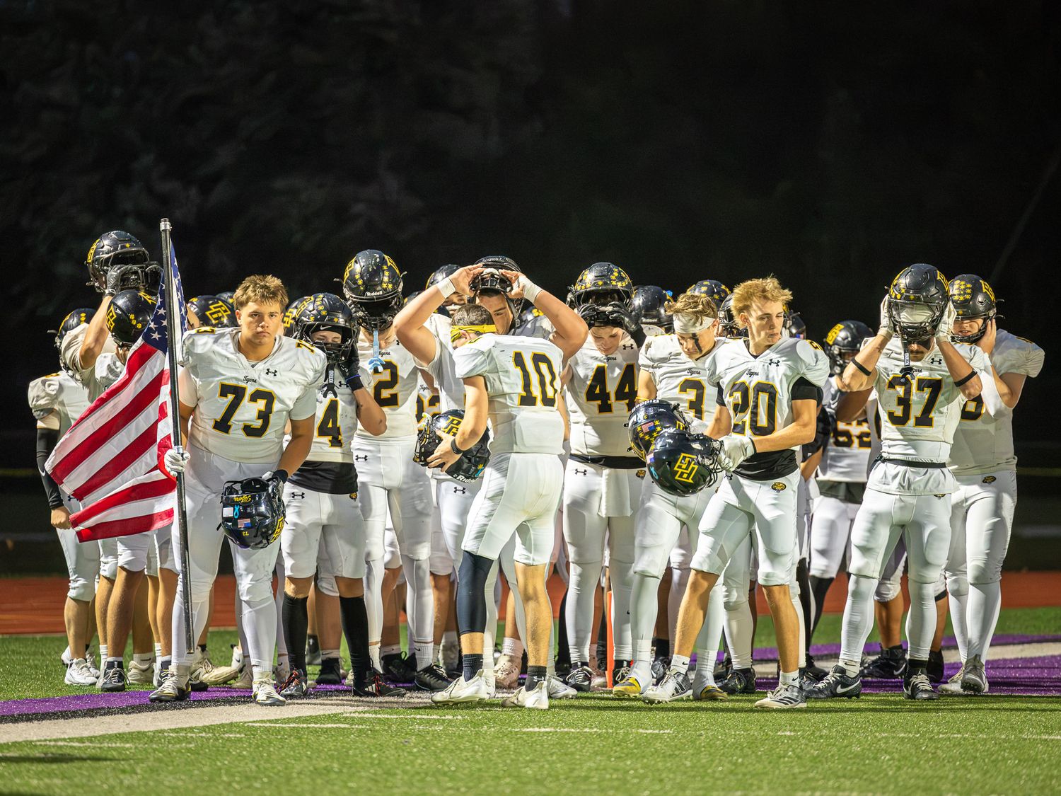 High school football players in white jerseys and helmets stand in a line on the field with an American flag during pregame ceremonies.