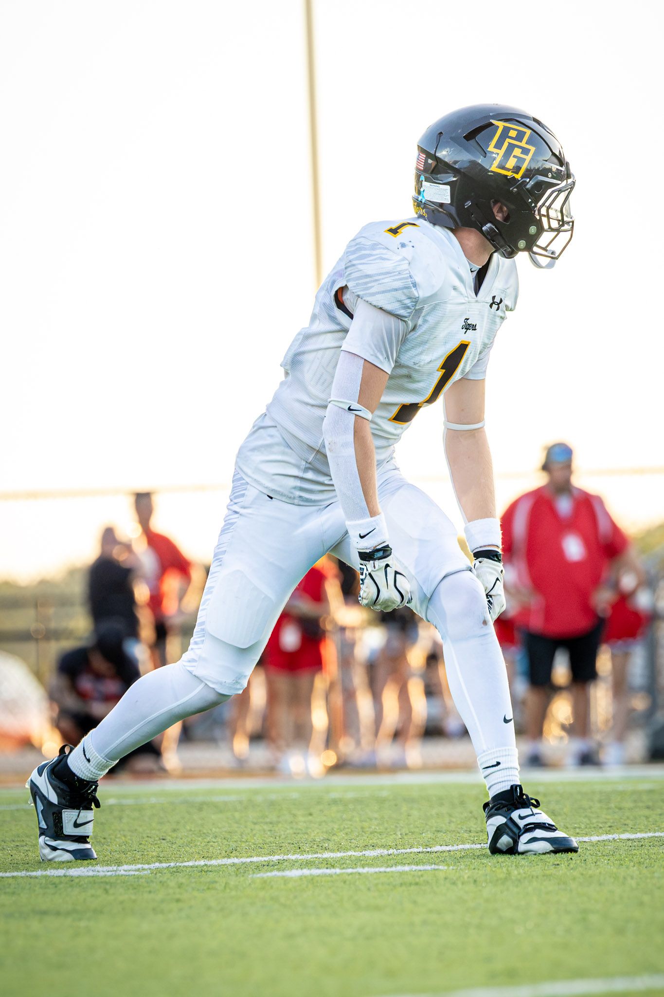 High school football player in a white jersey, black and gold helmet, and gloves crouches in a ready stance on the field during a game.