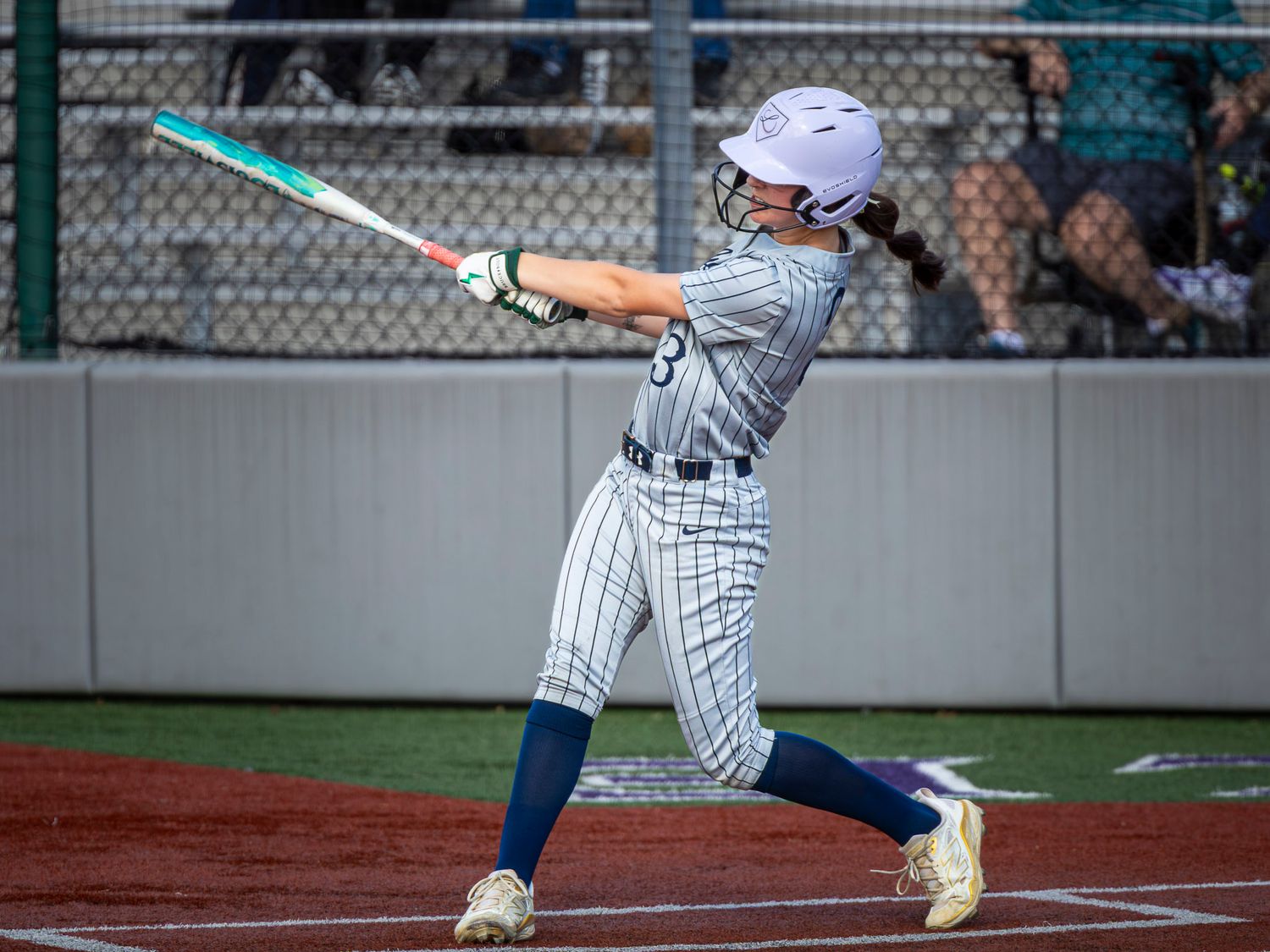 A softball batter in a white helmet and pinstriped uniform swings the bat through a pitch during a game.