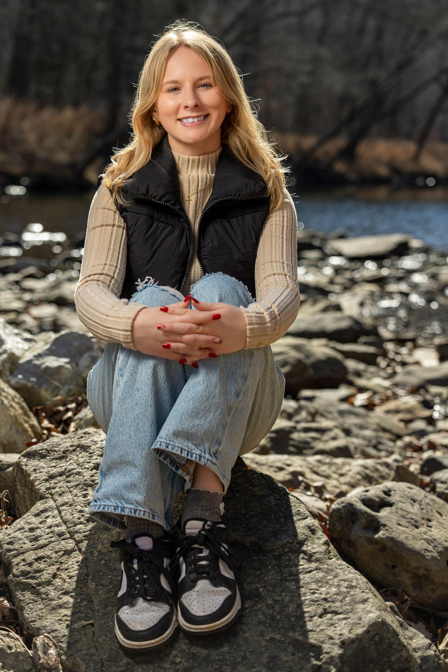 Smiling blonde woman sitting on rocks by a river, wearing a black vest, beige sweater, jeans, and sneakers.