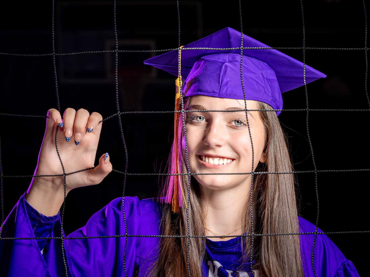 Smiling graduate in a purple cap, gown and tassel gripping a volleyball net while posing against a dark background.