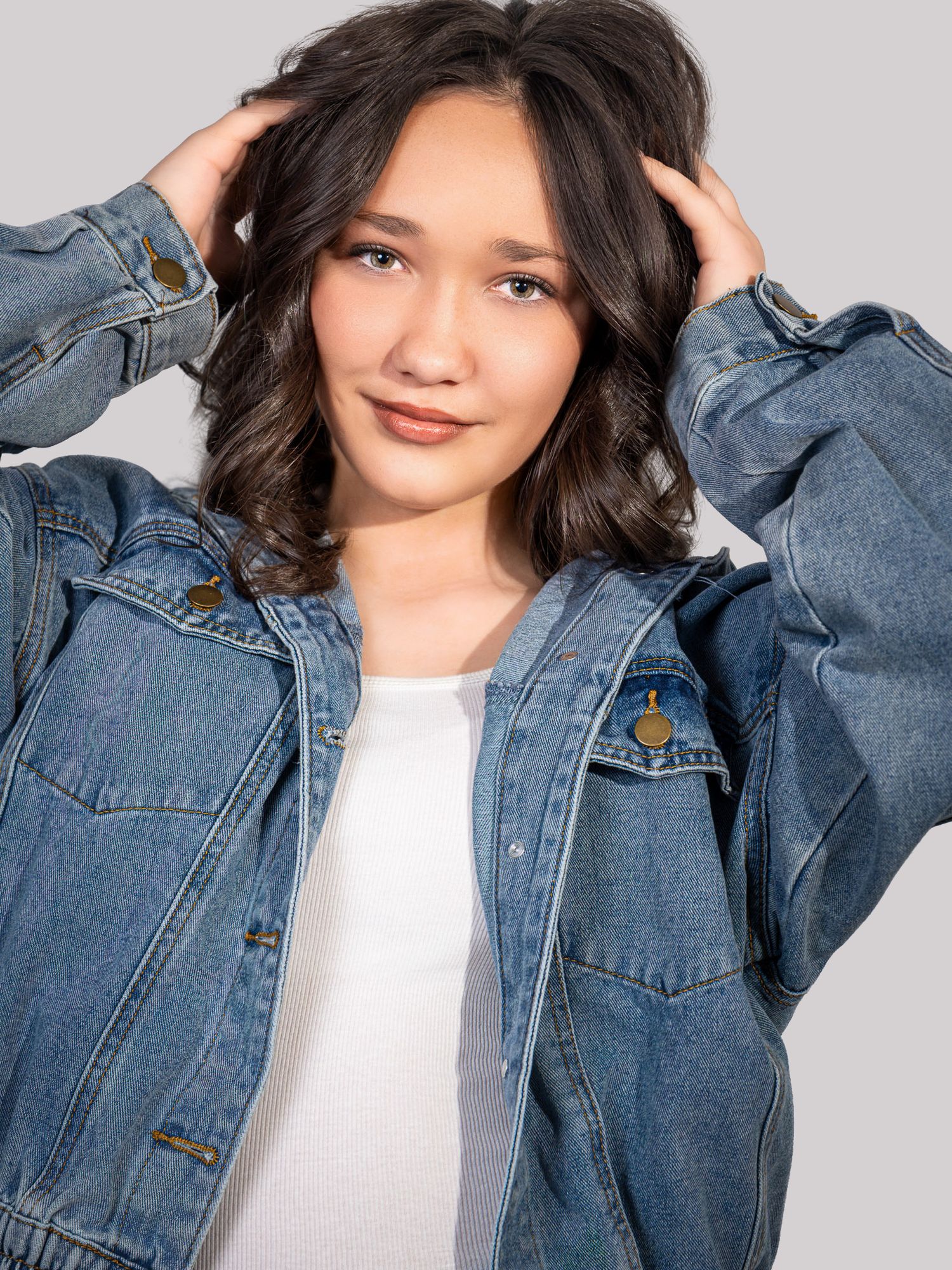 Young woman in oversized blue denim jacket over white top, hands in dark wavy hair, smiling against gray background.