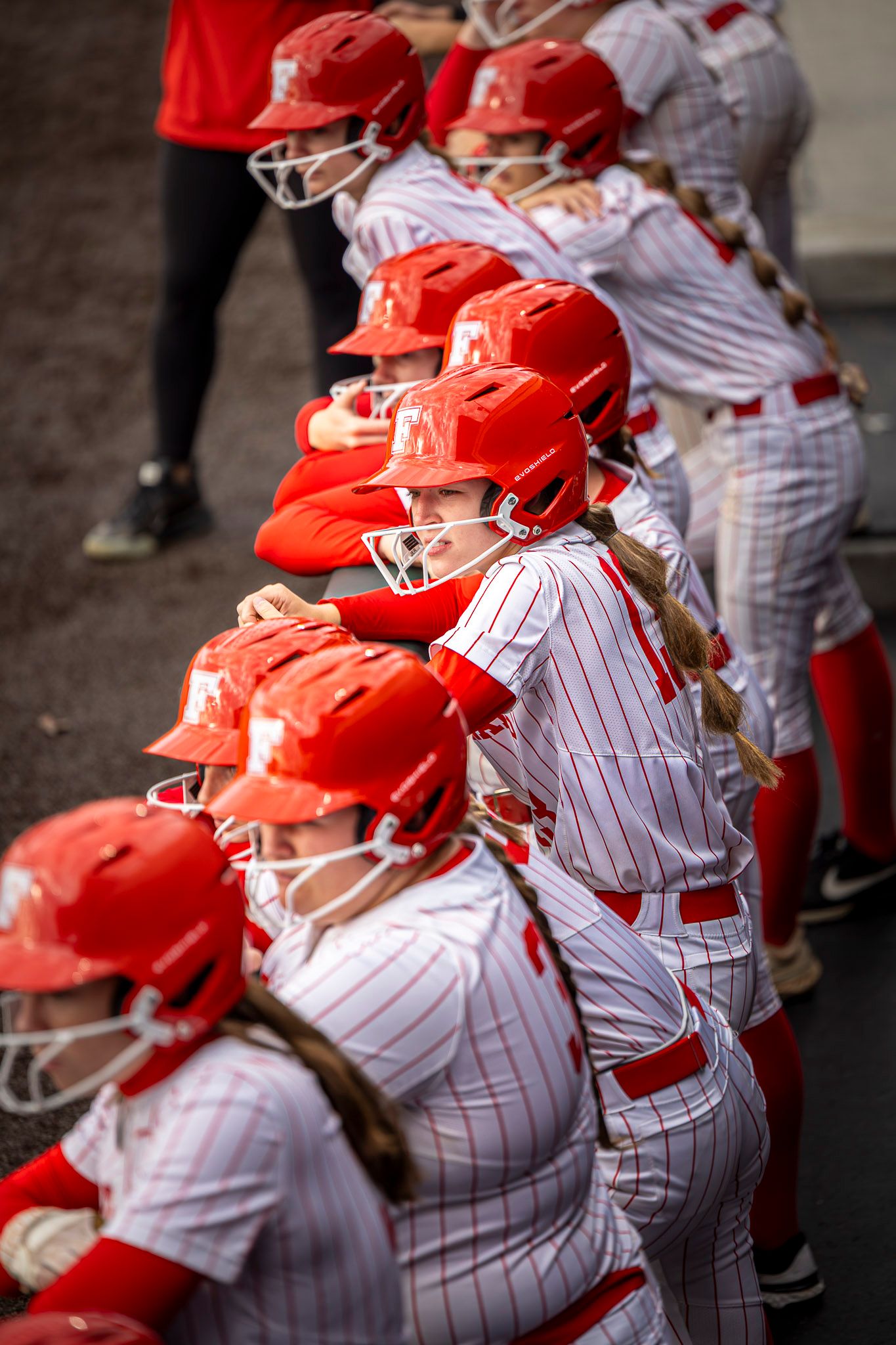 Softball teammates in red helmets with face guards and pinstripe uniforms lean on the dugout rail, watching the game.