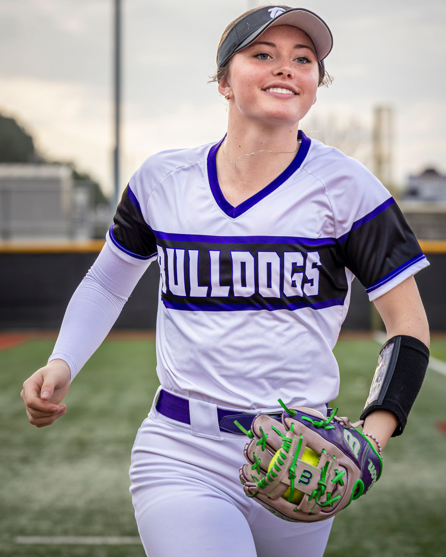 Smiling softball player in a white and purple Bulldogs jersey and gray visor holds a green and purple glove while walking across the field.