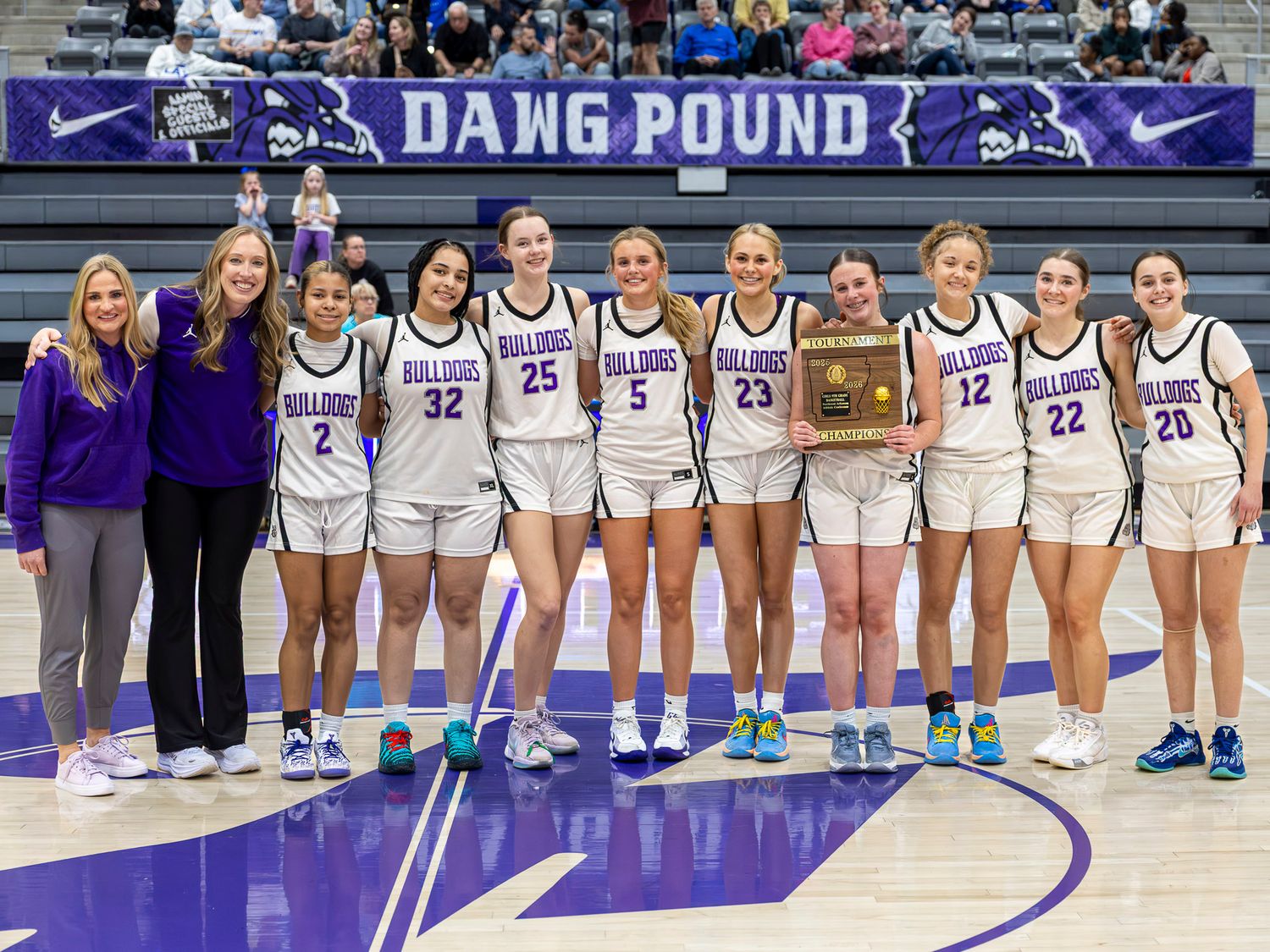 College basketball team poses together on court during game day wearing white and purple uniforms.