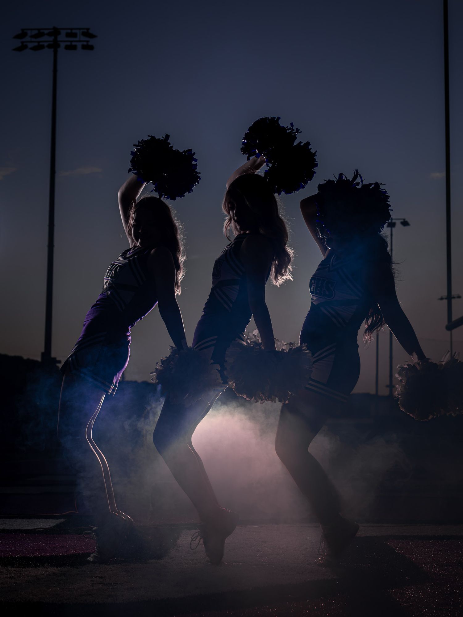 Silhouetted cheerleaders with pom-poms perform in dramatic fog and blue light at dusk on a football field.