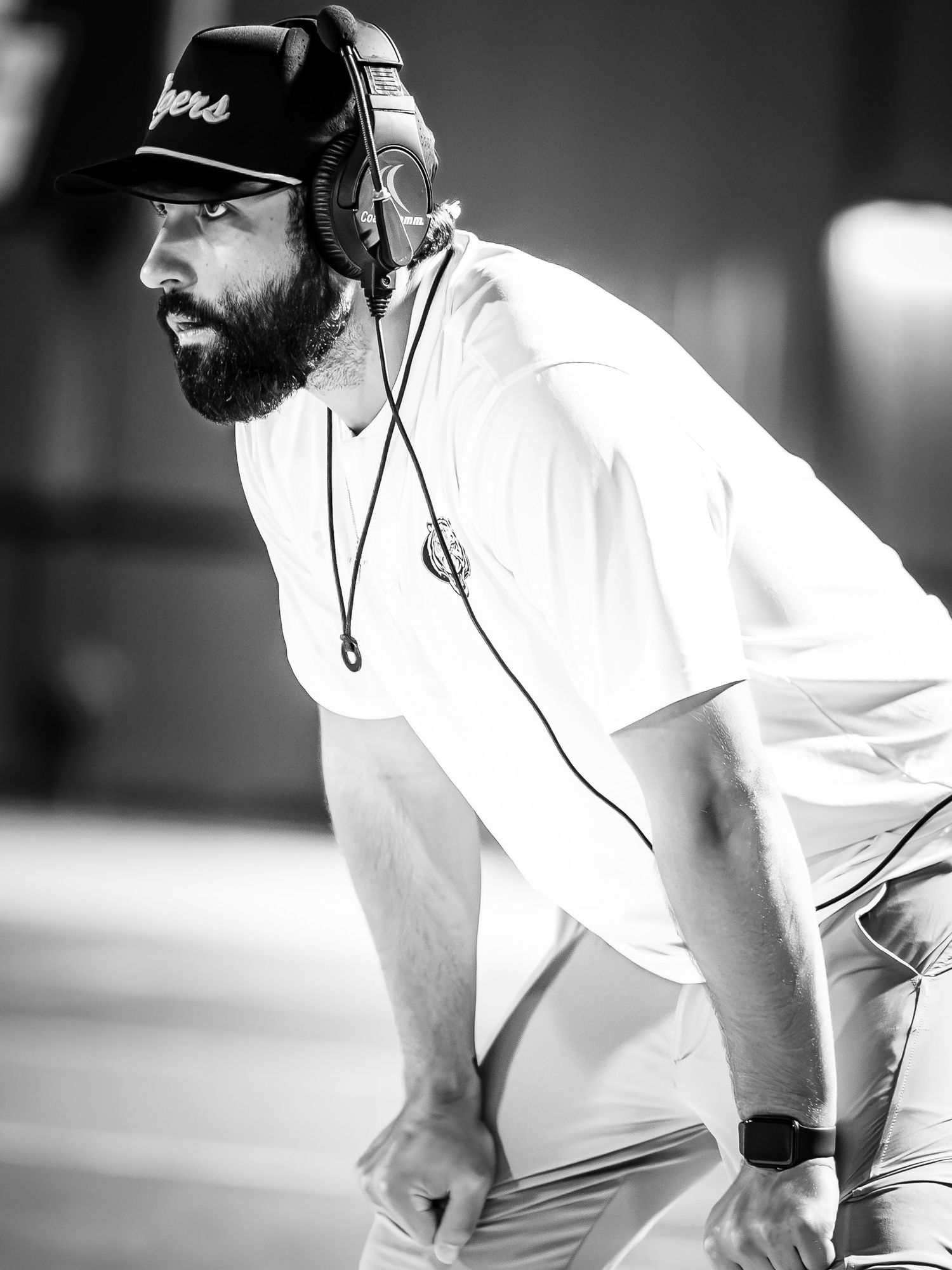 Black and white photo of a bearded coach in a cap and headset leaning forward with hands on knees, watching intently from the sideline.