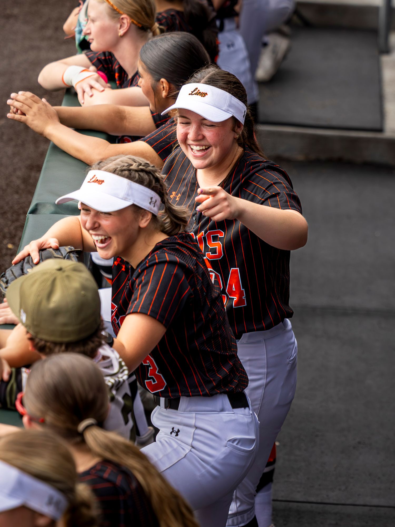 Smiling softball players in black pinstripe jerseys and white visors laugh and cheer together while leaning on the dugout railing.